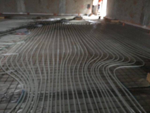 Zakřivené trubky nebo trubky jsou instalovány v paralelních řadách na kovové konstrukci ve tvaru mřížky na betonové podlaze, pravděpodobně pro systém podlahového vytápění, ve velké nedokončené budově.