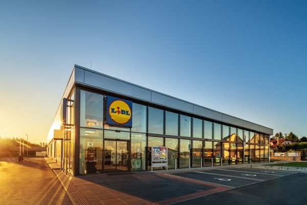 Moderní supermarket Lidl s velkými prosklenými okny a logem Lidl na budově, na snímku při západu slunce s jasnou modrou oblohou a prázdným parkovištěm před ním.